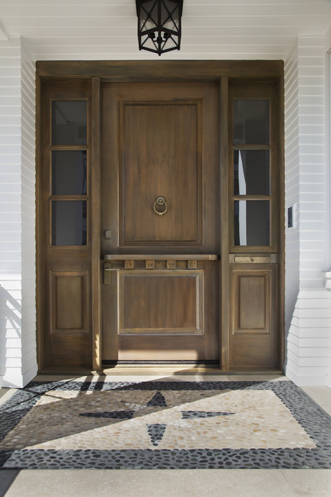 Vertical shot of a tall wooden front door on an upscale home with view of windows and decorative floor tiling