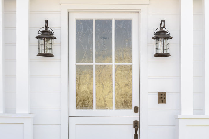 Cropped horizontal shot of a white door to a classic, white, family home in daytime. The door has intricate, etched, glass detail. Also see is light fixtures, columns, and a door bell.